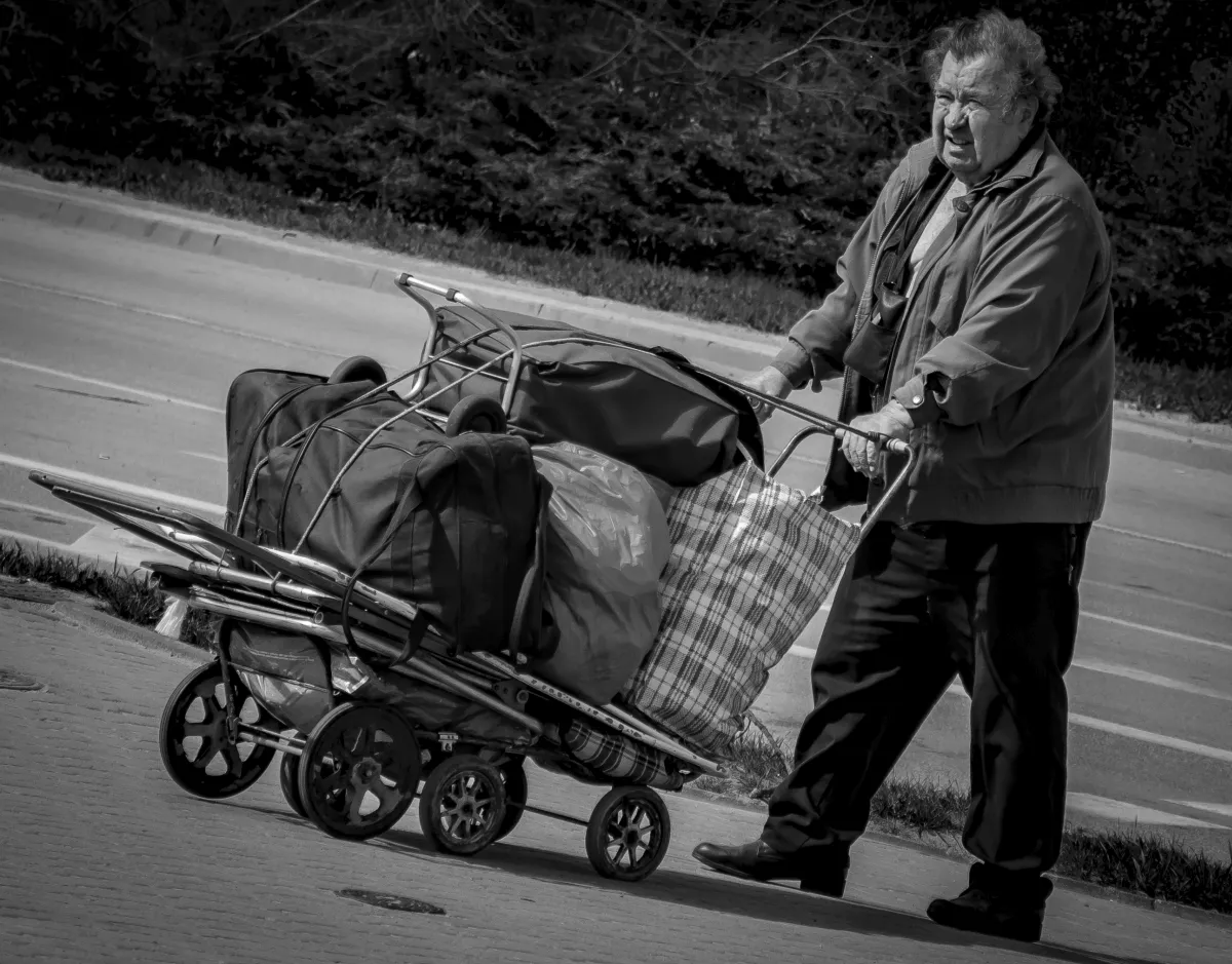 Older man pushing an overloaded trolley along a roadside sidewalk